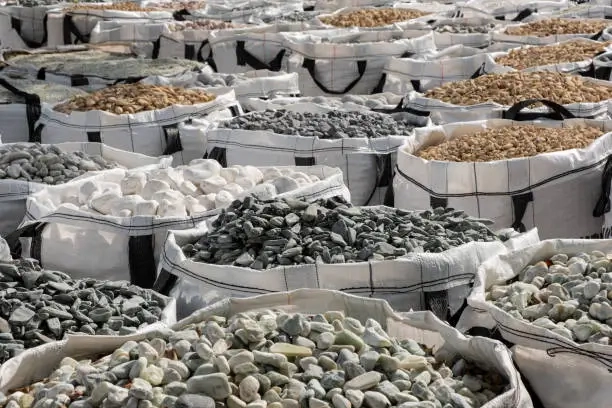 Large fabric bags filled with different types and colors of decorative landscaping stones, including white, gray, tan, and green stones, displayed at a garden supply or stone yard.